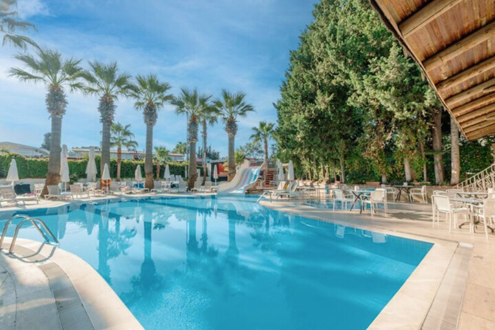 Hotelpool mit Wasserrutsche, Palmen und mehreren Sonnenliegen unter Sonnenschirmen, daneben Tische und Stühle auf der Terrasse.