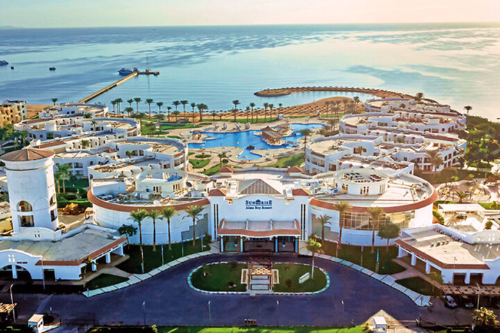 Luftaufnahme eines weißen Hotelkomplexes mit runden Gebäuden und Palmen an einer Bucht bei Sonnenaufgang.