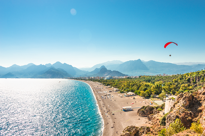 Ein Paraglider fliegt über die Küste, mit majestätischen Bergen im Hintergrund und dem glitzernden Meer darunter.