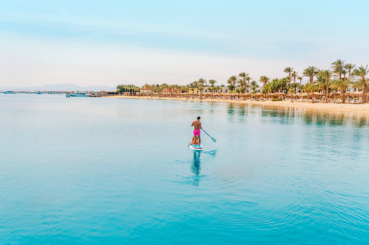 Person auf einem Stand-up-Paddle-Board auf ruhigem, türkisfarbenem Wasser vor einer Küste mit Palmen und Gebäuden.