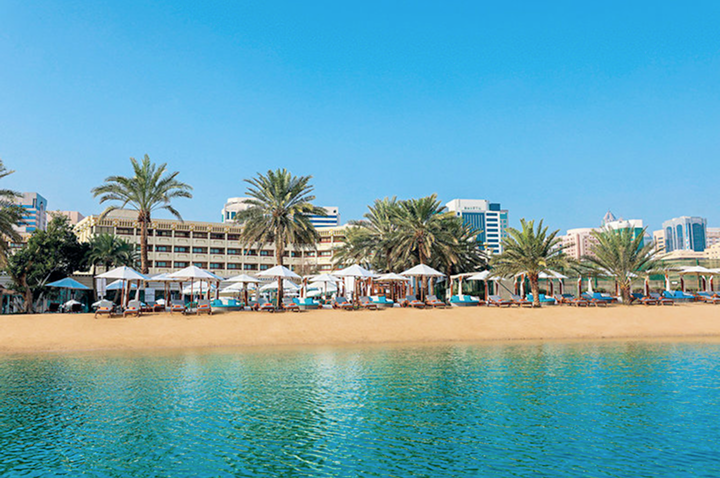 Ein Strand mit einem Hotel und einigen Palmen, die sanft im Wind wiegen, unter einem strahlend blauen Himmel.