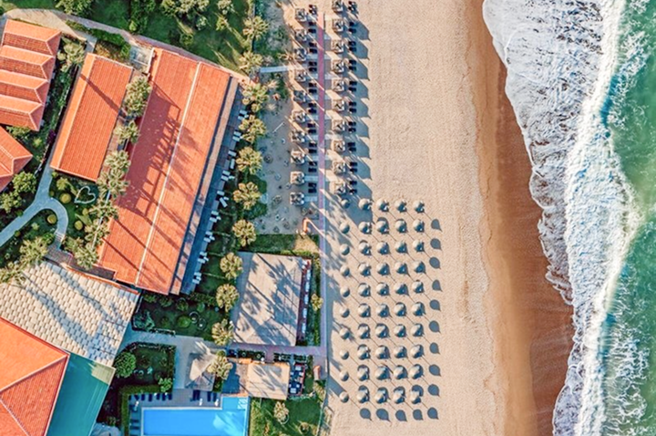 Luftaufnahme eines Strandes mit Reihen von Sonnenschirmen und Liegen neben einem Hotel mit roten Dächern und einem Pool.