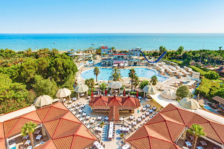 Luftaufnahme einer weitläufigen Hotelanlage mit mehreren roten Dächern, Poollandschaft, Palmen und Meer im Hintergrund.