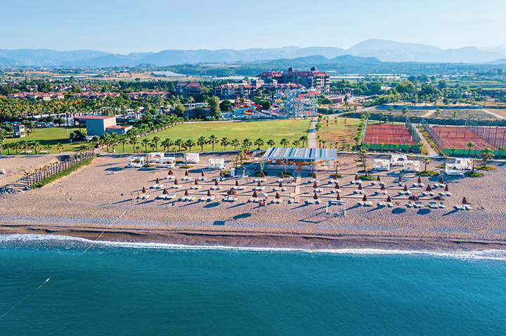 Luftaufnahme eines Strandes mit Sonnenschirmen, dahinter Grünflächen, Gebäude und Berge im Hintergrund