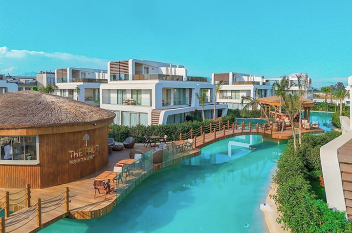 Moderner Hotelkomplex mit mehreren weißen Gebäuden, einem türkisfarbenen Wasserkanal und einem runden Restaurant mit Strohdach.