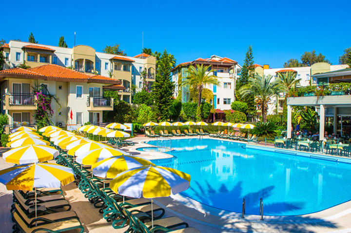 Großer Pool mit mehreren gelb-weißen Sonnenschirmen und Liegestühlen, umgeben von mehrstöckigen Gebäuden und Palmen.