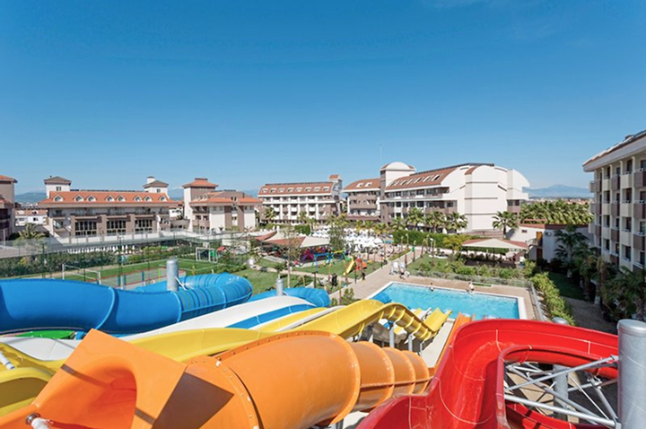 Mehrere bunte Wasserrutschen führen in ein Schwimmbecken vor einem Hotelkomplex mit mehreren Gebäuden unter blauem Himmel.