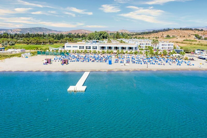Luftaufnahme eines Strandes mit einem Hotel, das direkt am Wasser liegt, umgeben von Palmen und Sonnenliegen.