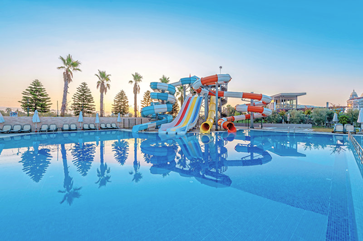 Wasserpark mit mehreren bunten Wasserrutschen und großem Schwimmbecken, umgeben von Liegestühlen und Palmen bei Sonnenuntergang. Wasserpark mit mehreren bunten Wasserrutschen und großem Schwimmbecken, umgeben von Liegestühlen und Palmen bei Sonnenuntergang.