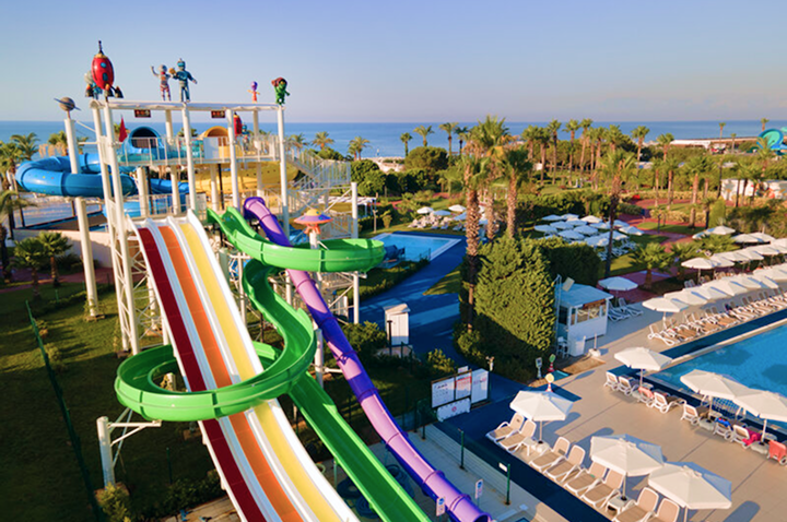 Wasserpark mit bunten Wasserrutschen, Liegestühlen und Sonnenschirmen neben einem Pool, umgeben von Palmen und Blick aufs Meer.
