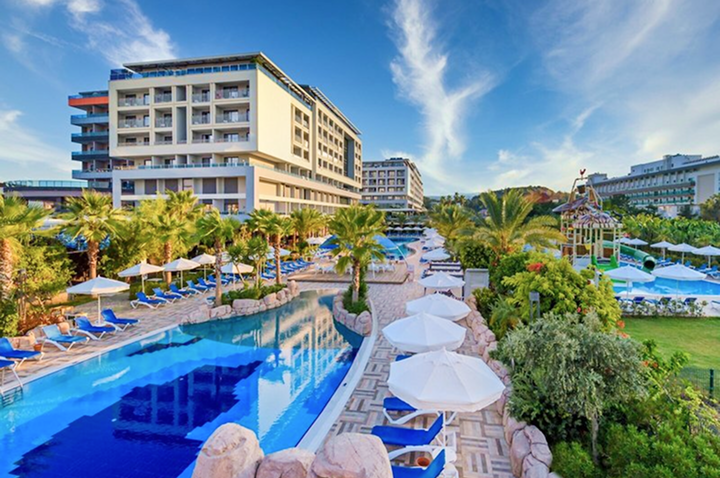 Hotelanlage mit mehreren mehrstöckigen Gebäuden, großem Pool, Liegen mit weißen Sonnenschirmen und Palmen unter blauem Himmel.
