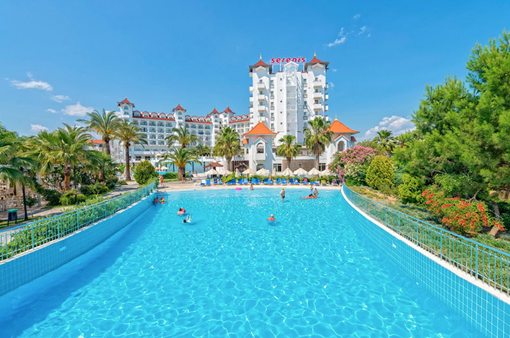 Hotelgebäude mit mehreren Stockwerken und roten Dächern hinter großem Schwimmbecken mit Menschen und umgeben von Palmen und Gartenanlagen.