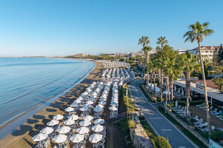 Strand mit Reihen von Sonnenschirmen und Liegestühlen neben einer Promenade mit Palmen und Gebäuden im Hintergrund.