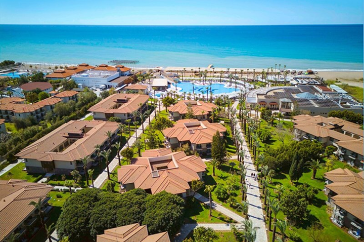 Luftaufnahme eines Ferienresorts mit mehreren Gebäuden, Pool, Palmen und Strand im Hintergrund.