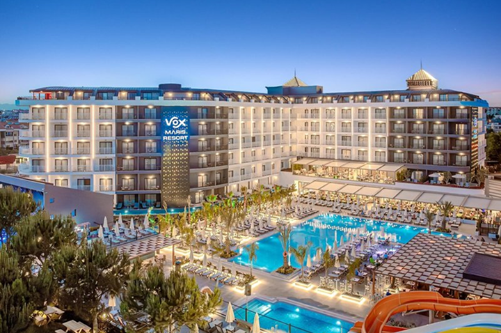 Großes Hotelgebäude mit beleuchtetem Schriftzug "VOX Maris Resort" und mehreren Pools mit Liegen und Sonnenschirmen davor.