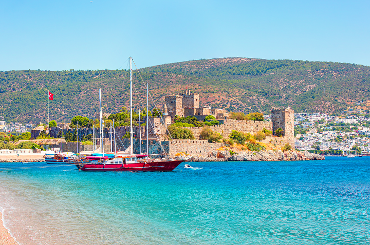 Burg mit mehreren Türmen an der Küste, davor Segelboote im klaren blauen Wasser, im Hintergrund bewaldete Hügel.