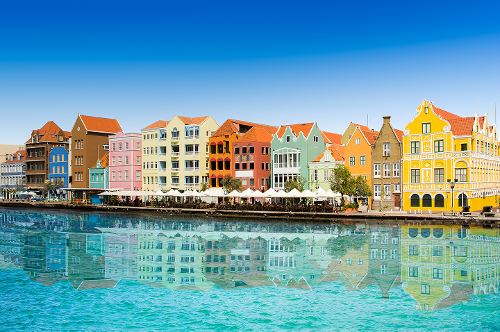 Reihe bunter, historischer Häuser am Wasser mit klarer Spiegelung im ruhigen Wasser unter blauem Himmel.