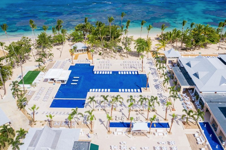 Luftaufnahme eines großen rechteckigen Pools mit Liegestühlen und Palmen, dahinter türkisfarbenes Meer und Strand.