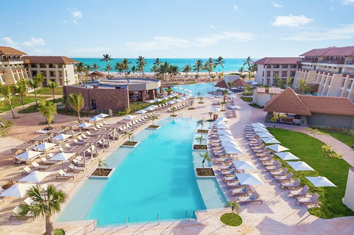 Großer Hotelpool mit Liegen und Sonnenschirmen, umgeben von mehrstöckigen Hotelgebäuden und Palmen, im Hintergrund das Meer.