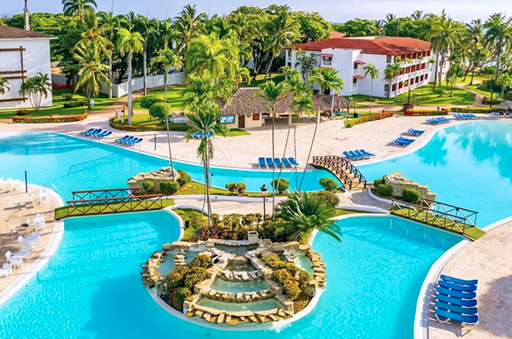 Luftaufnahme einer Hotelanlage mit mehreren Pools, Palmen, Liegen und weißen Gebäuden mit roten Dächern.