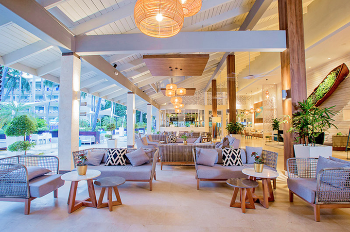 Offener, heller Lounge-Bereich mit modernen Sitzmöbeln, Holztischen, Pflanzen und hängenden Lampen unter einem weißen Satteldach.