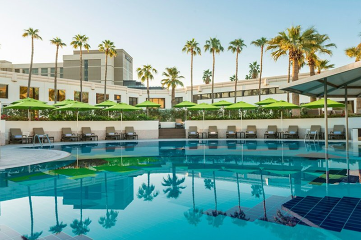 Hotelgebäude mit mehreren Stockwerken hinter einem Pool mit Liegestühlen und grünen Sonnenschirmen, umgeben von Palmen. Hotelgebäude mit mehreren Stockwerken hinter einem Pool mit Liegestühlen und grünen Sonnenschirmen, umgeben von Palmen.