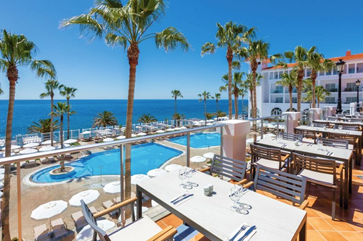 Terrasse mit gedeckten Tischen und Stühlen neben einem Pool, umgeben von Palmen mit Blick auf das Meer und ein weißes Hotelgebäude im Hintergrund.
