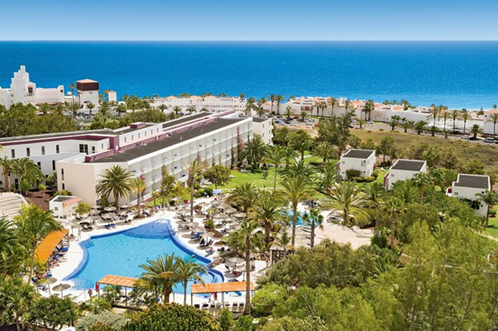 Großer Hotelkomplex mit braunen Dächern, mehreren Stockwerken und einem Pool mit blauer Wasserfläche, umgeben von Liegestühlen und Sonnenschirmen.