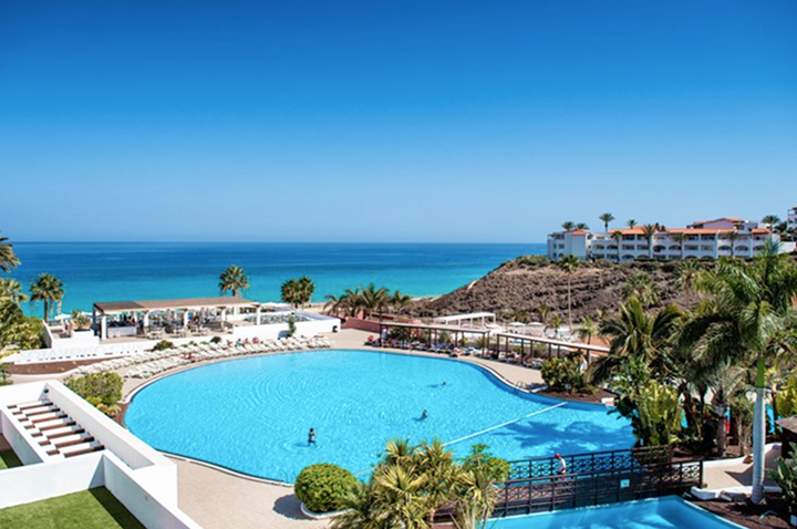 Großer, rundlicher Pool mit wenigen Personen, umgeben von Palmen und weißen Gebäuden, im Hintergrund das blaue Meer unter klarem Himmel.