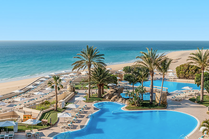 Großer Pool mit Palmen und Liegestühlen an einem Sandstrand mit Blick auf das Meer.