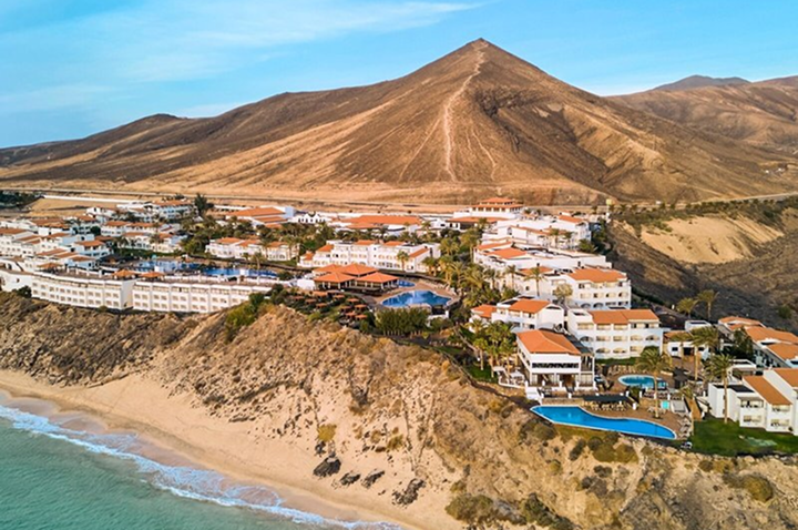 Hotelanlage mit mehreren Gebäuden und Pools auf einer Klippe am Meer vor einem kahlen Berg.