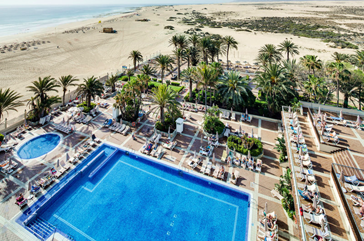 Großer rechteckiger Pool mit Liegestühlen und Palmen neben einem Sandstrand