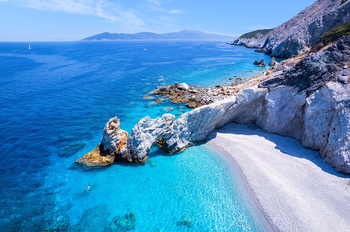 Klippen mit Felsenbogen über türkisfarbenem Meer und Kiesstrand an einer Küste mit Bergen im Hintergrund.