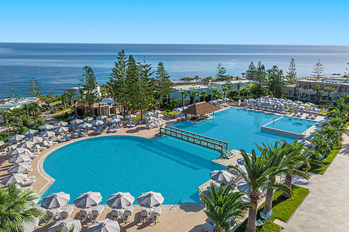 Großer Hotelpool mit Liegestühlen und Sonnenschirmen am Meer und umgeben von Palmen und Gebäuden