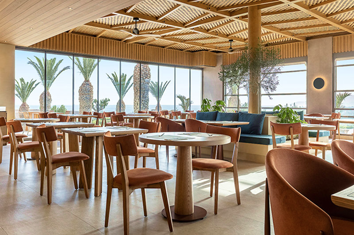 Helles Restaurant mit Holzbalkendecke, großen Fenstern, Palmenblick, Tischen mit braunen Stühlen und blauen Sitzbänken. Helles Restaurant mit Holzbalkendecke, großen Fenstern, Palmenblick, Tischen mit braunen Stühlen und blauen Sitzbänken.