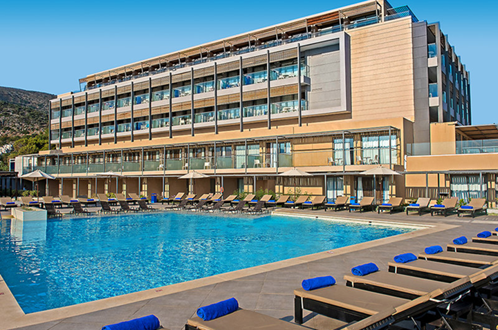 Modernes Hotelgebäude mit mehreren Etagen und großen Glasfronten hinter einem rechteckigen Außenpool, umgeben von Liegen mit blauen Auflagen.