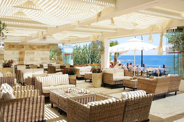 Terrassenbereich mit geflochtenen Sitzmöbeln und weißen Polstern unter einem Sonnenschutz, im Hintergrund Meer und Sonnenschirme.