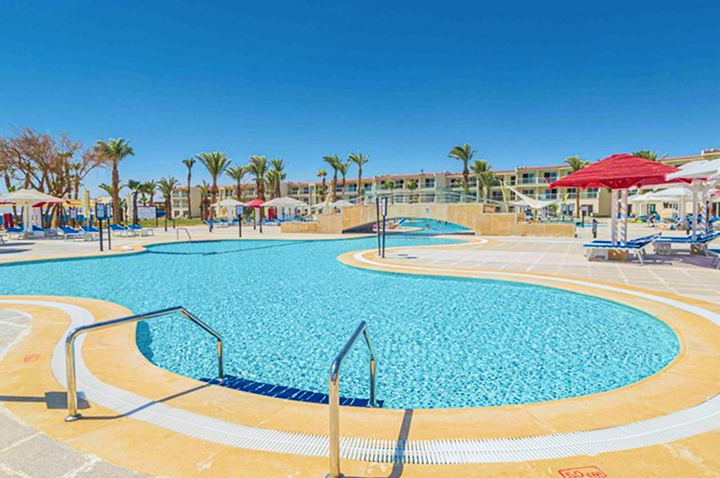 Großer, geschwungener Pool mit Edelstahlgeländern, umgeben von Liegestühlen, Sonnenschirmen und Palmen vor einem mehrstöckigen Hotel.