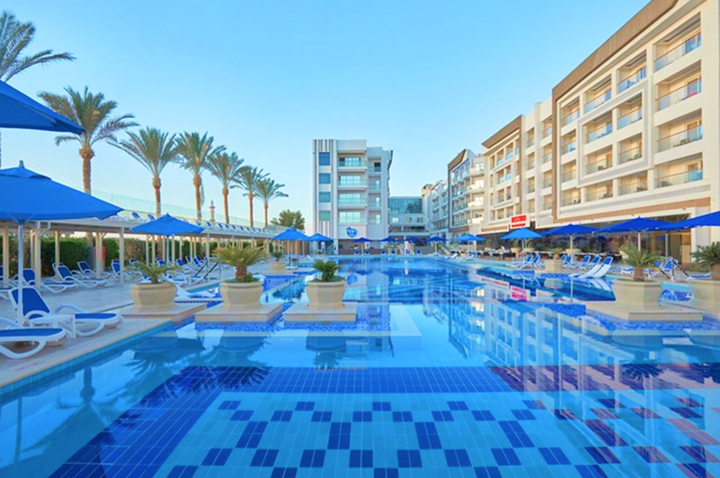 Großer rechteckiger Pool mit blauen Sonnenschirmen und Liegestühlen, umgeben von mehrstöckigen Hotelgebäuden und Palmen.