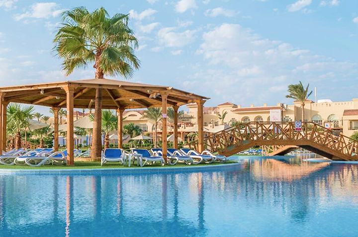 Pool mit Liegestühlen unter einem Holzpavillon und einer Holzbrücke vor einem Hotelgebäude mit Palmen.