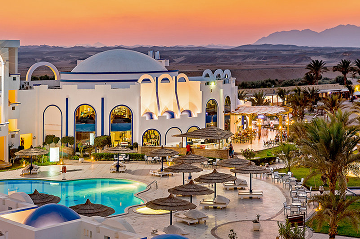 Hotelanlage mit weißer, kuppelförmiger Architektur, Pool und mehreren Sonnenschirmen bei Sonnenuntergang in einer Wüstenlandschaft.