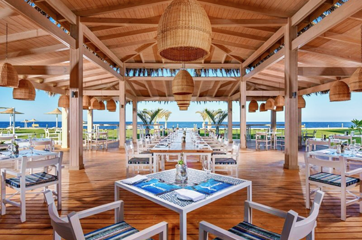 Offener Restaurantbereich mit Holzdach, mehreren Tischen und Stühlen, Blick auf Meer und Palmen.