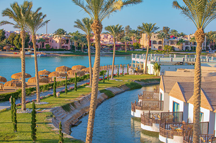 Bungalows mit Holzterrassen am Wasser, umgeben von Palmen und Grünflächen, im Hintergrund weitere Gebäude.