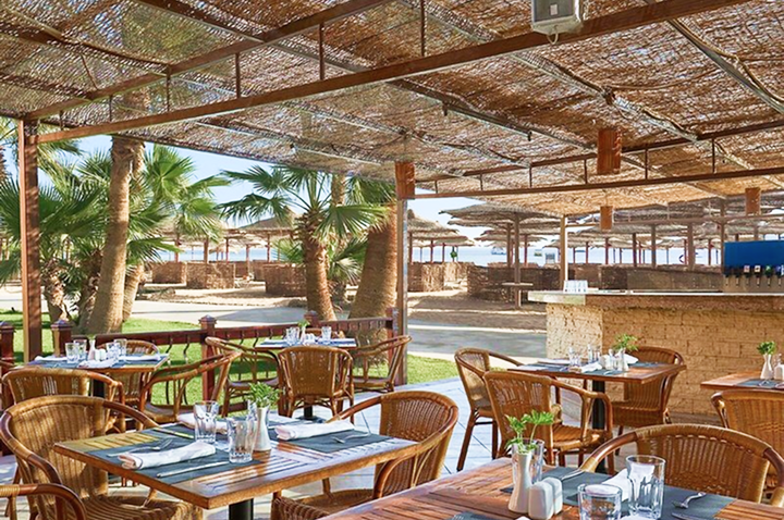 Überdachte Terrasse mit Holztischen und Korbstühlen vor einer Gartenanlage mit Palmen und Sonnenschirmen.