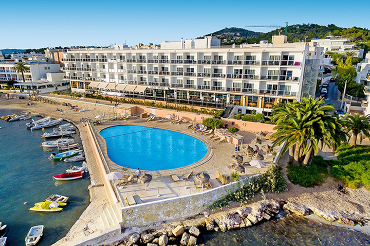 Hotelgebäude mit mehreren Stockwerken und Balkonen, davor ein rundlicher Pool, umgeben von Liegestühlen und Palmen, angrenzend an eine kleine Hafenanlage mit Booten.