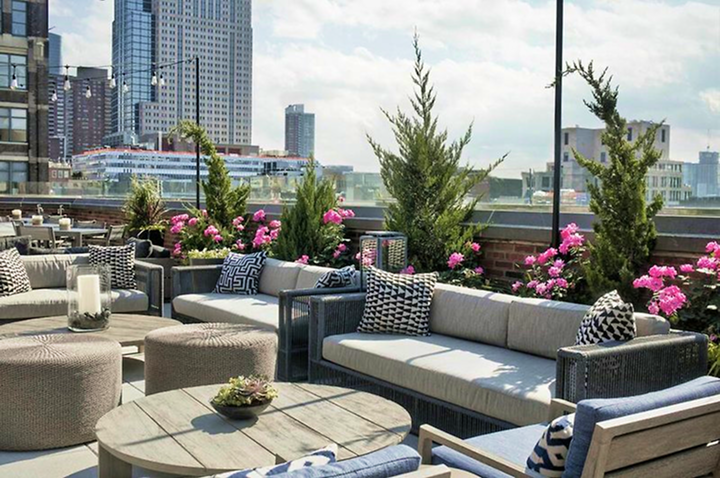 Dachterrasse mit modernen Sitzmöbeln aus Rattan, grauen Polstern, runden Hockern, Holztisch und blühenden pinken Blumen vor Stadtkulisse.