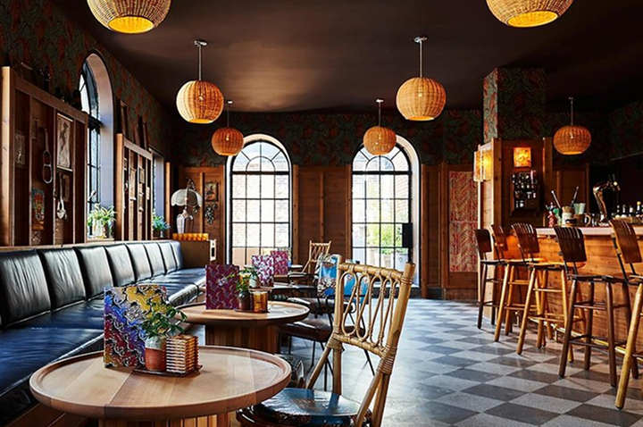Hotelbar mit runden Holztischen, Stühlen aus Holz und Rattan, Ledersitzbank, großen Bogenfenstern und hängenden Lampen mit warmem Licht.