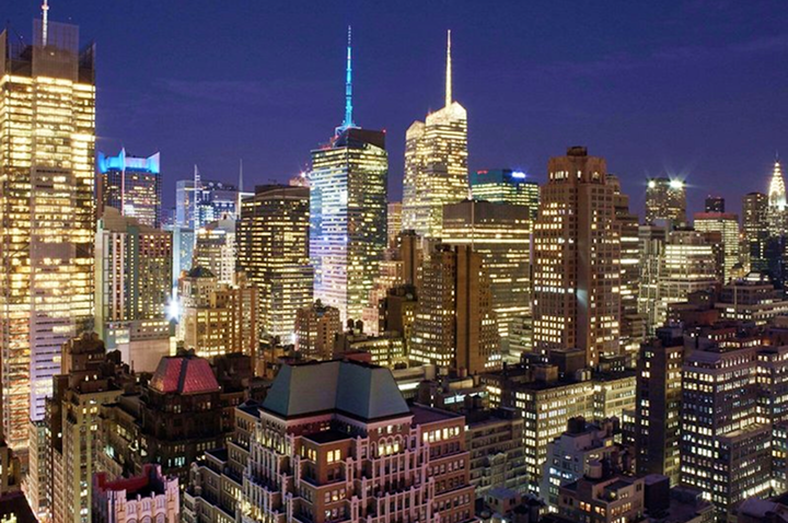 Dachterrasse mit modernen Sitzmöbeln aus Rattan, grauen Polstern, runden Hockern, Holztisch und blühenden pinken Blumen vor Stadtkulisse.