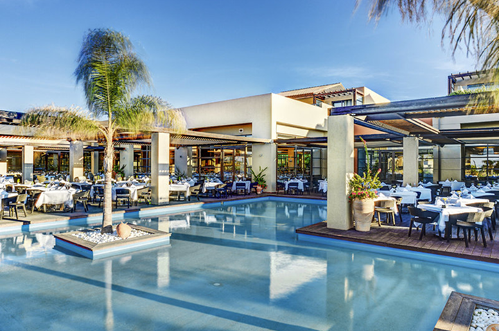 Außenbereich eines Restaurants mit Pool, Tischen und Stühlen unter Sonnendächern und Palmen.