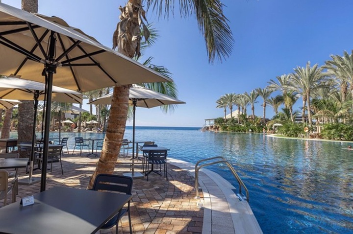 Poolbereich mit Sonnenschirmen, Tischen und Stühlen neben einem großen Schwimmbecken und Palmen im Hintergrund.